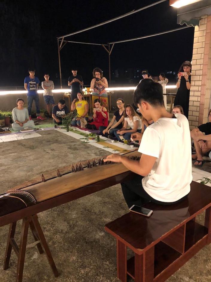 a man in a white shirt plays the chinese guzheng instrument for a small audience on a rooftop at night