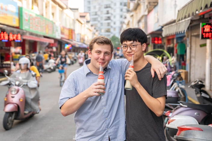 a Chinese and western boy stand outside sipping a drink with their arms around each other