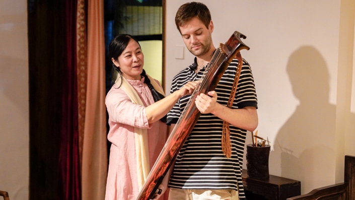 a student holding an ancient Chinese instrument while his teacher looks on