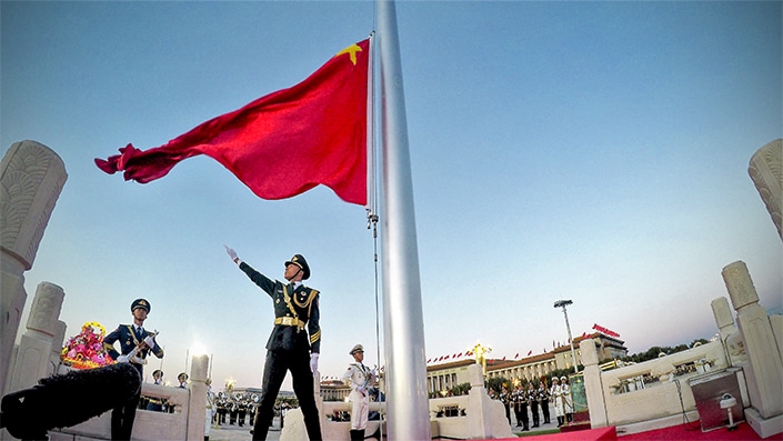 Un soldado chino en uniforme hace gestos con una mano debajo de una bandera china roja durante la ceremonia de izamiento de la bandera del Día Nacional de China.