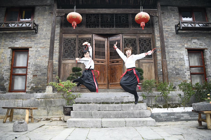 two women holding traditional martial arts weapons jumping into the air in front of a traditional Chinese house with red lanterns