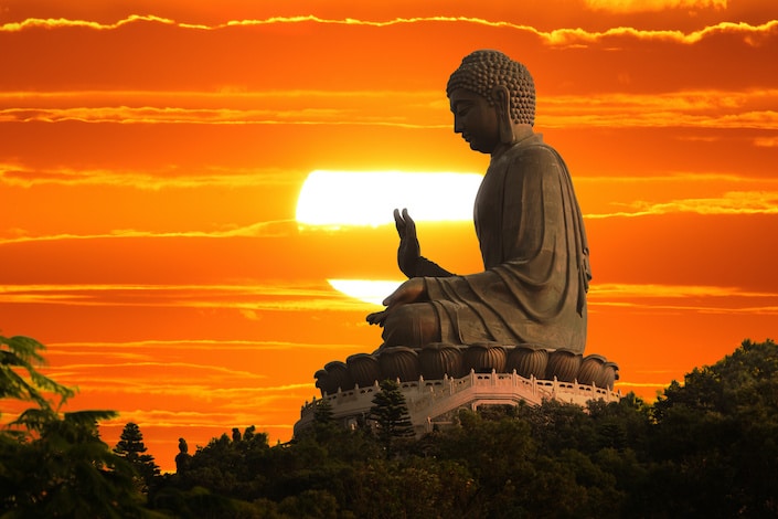 a Buddha statue with one hand raised against a setting sun and orange sky