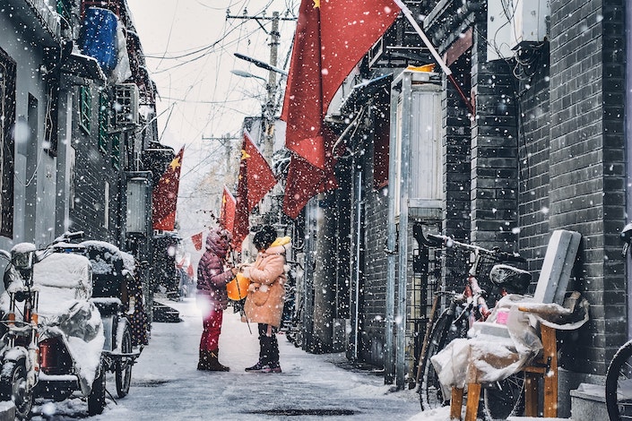 a snowy winter scene showing two people in winter coats standing in an alley with traditional Chinese grey brick buildings and red Chinese flags on either side