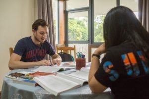 A student reads a textbook at a round table while a teacher looks on