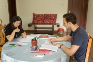 Two students write in workbooks at a round table covered with books