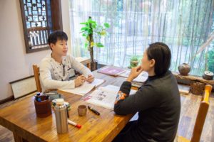 A student and teacher talk across a wooden table by a bright window
