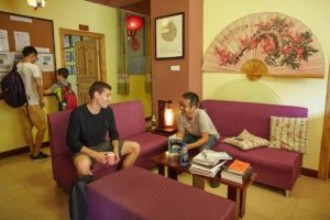 Two men chat on maroon sofas while others read a notice board