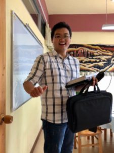 A smiling man with a laptop bag stands by a classroom whiteboard