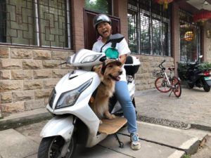 A smiling woman rides a scooter with a dog on the footboard