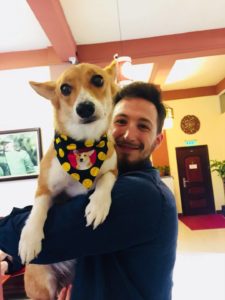 A smiling man holds a shiba inu wearing a black and yellow bandana