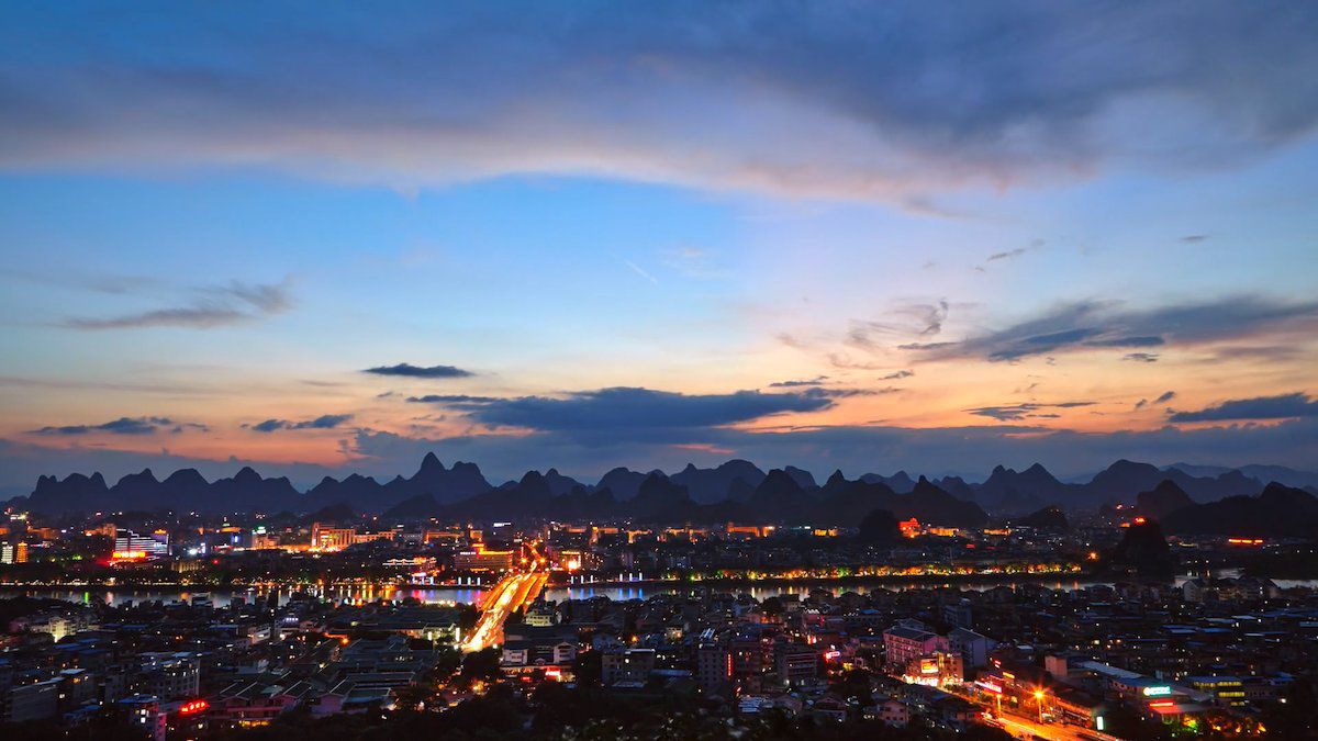 a photo of a bridge and buildings in downtown Guilin illuminated with lights after sunset with shadowy karst mountains in the background
