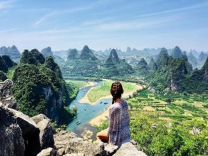 A woman sits on a cliff overlooking a winding river and karst peaks