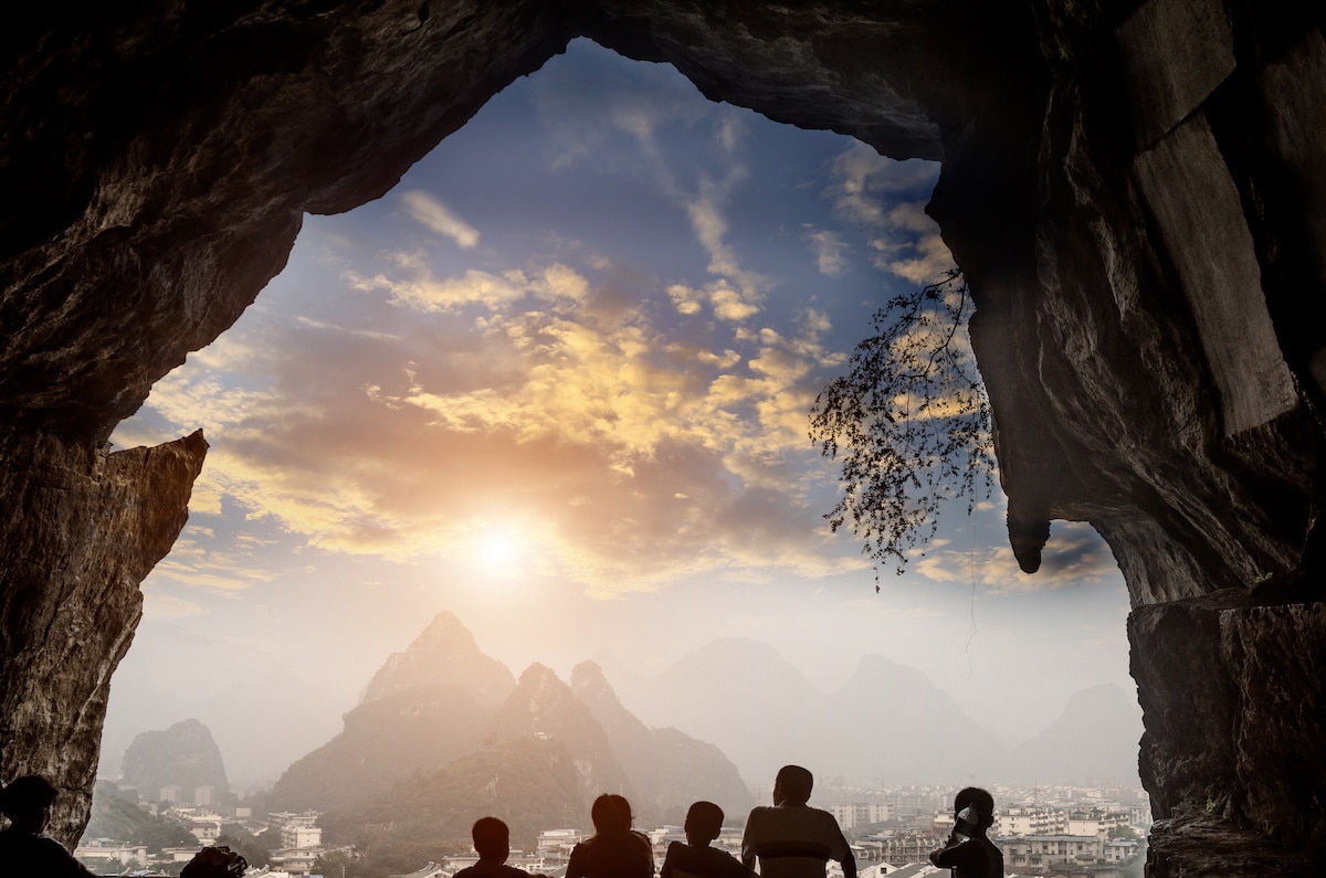 Five people silhouetted against the sky staring out at karst mountains with the setting sun in front of them framed by the mouth of a cave