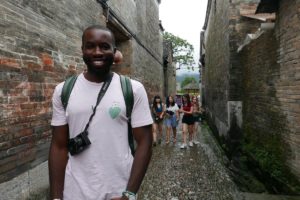 A smiling man with a camera stands in a narrow old village lane