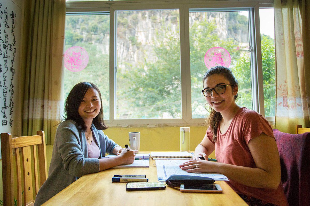 在中国桂林，一位戴着大眼镜的西方女性和她的中文老师坐在一张桌子旁，身后有一扇窗户