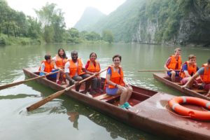 People in orange life jackets sit in two wooden rowboats on a river below cliffs
