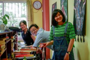 Three people smile in a busy office stacked with books
