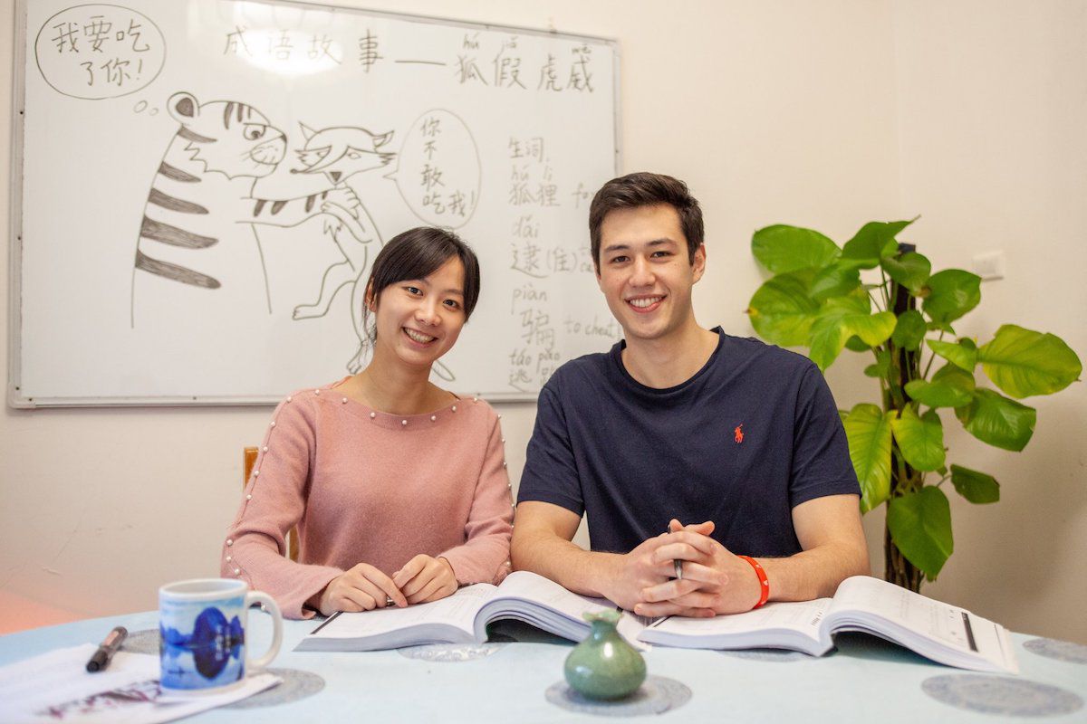 una maestra de chino y su estudiante occidental se sientan en una mesa durante la clase con una pizarra con un dibujo y caracteres chinos detrás de ellos