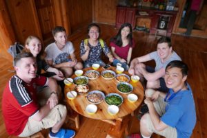 A group sits on the floor around a low round table of shared dishes