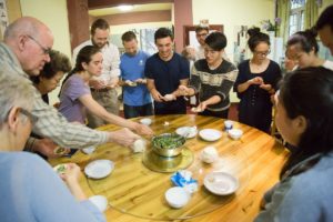 A large group folds dumplings around a round glass-topped table