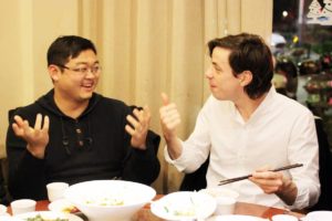 Two men talk and laugh over dinner with chopsticks in hand