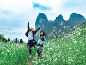 Two people jump in a field of white flowers below karst peaks