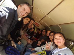 A large group squeezes onto benches for a selfie under a covered vehicle roof