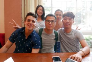 Five friends smile and make peace signs at a table by a window