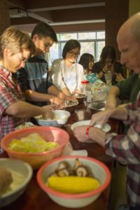 A group prepares food beside bowls of corn and mushrooms at a crowded table