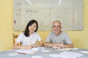 A teacher and student smile with pens in hand below a whiteboard of characters