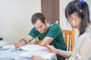 A smiling student writes in a workbook beside his teacher