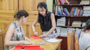 Two women work on red paper cuttings at a table by bookshelves