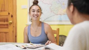 A smiling student in a tank top faces her teacher across a table of open books