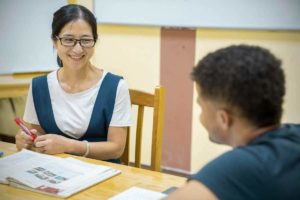 A teacher in glasses and a blue pinafore smiles at a student across the table