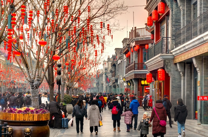 a winter street scene in China with red lanterns handing from trees and traditional Chinese buildings as people walk down the street