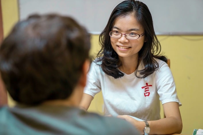 안경을 쓰고 흰색 티셔츠에 CLI 로고가 있는 웃고 있는 중국 여성이 맞은편에 앉아 있는 학생을 바라보고 있습니다.