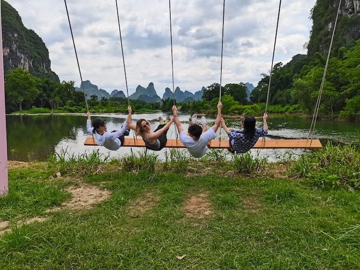 a group of four people sitting on a five-person swing and swinging towards a lake with karst mountains in the background