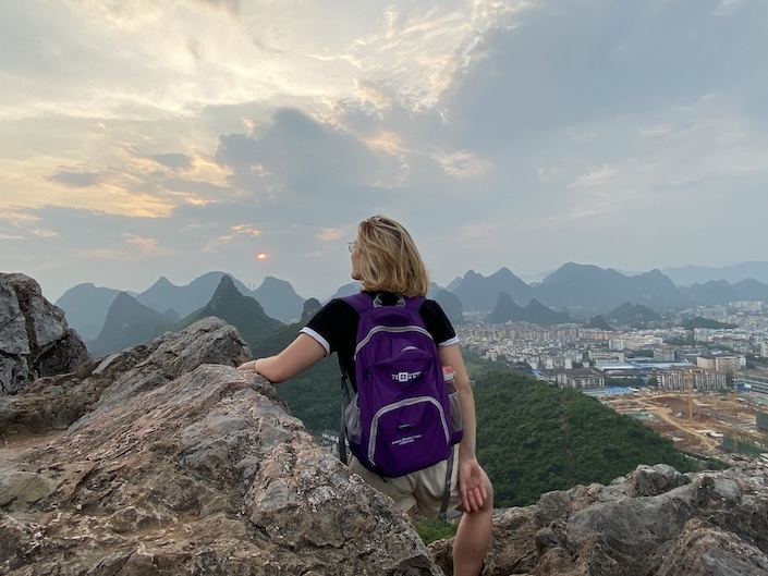 a blonde woman wearing a purple backpack sitting on top of a rock on a mountain overlooking karst peaks in Guilin, China