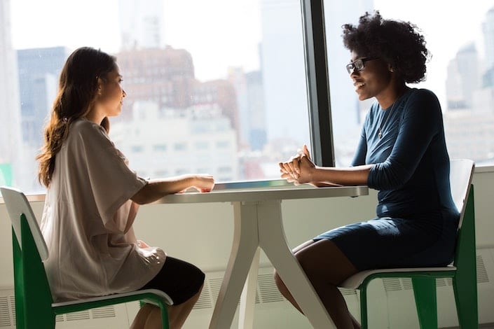 a Chinese woman and an African American woman sit at a table facing each other in front of a window