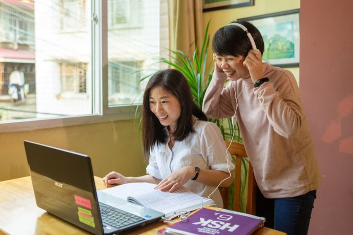 한 명은 앉고 한 명은 헤드폰을 끼고 서 있는 두 명의 중국 여성이 탁자 위의 노트북 컴퓨터를 보고 있다