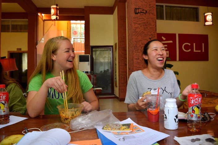 Una mujer occidental rubia que sostiene los palillos ríe mientras está sentada junto a una mujer china que se ríe con el logotipo de CLI en el fondo