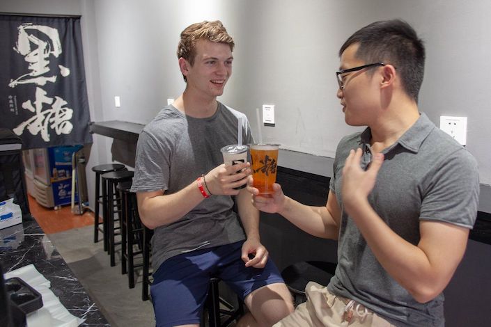 a young blonde man and a young Chinese man toasting each other with plastic cups full of liquid in a restaurant