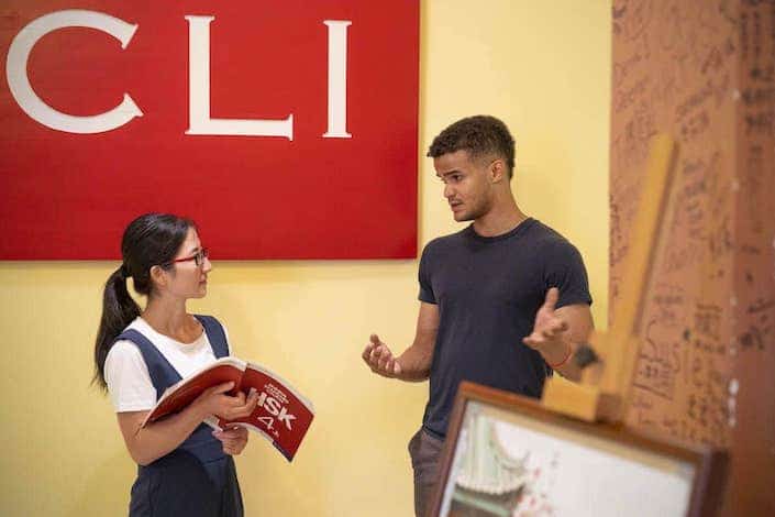 一名非洲裔美国男子在 CLI 标志前与一名手持 HSK 4 教科书的中国女教师交谈时做手势