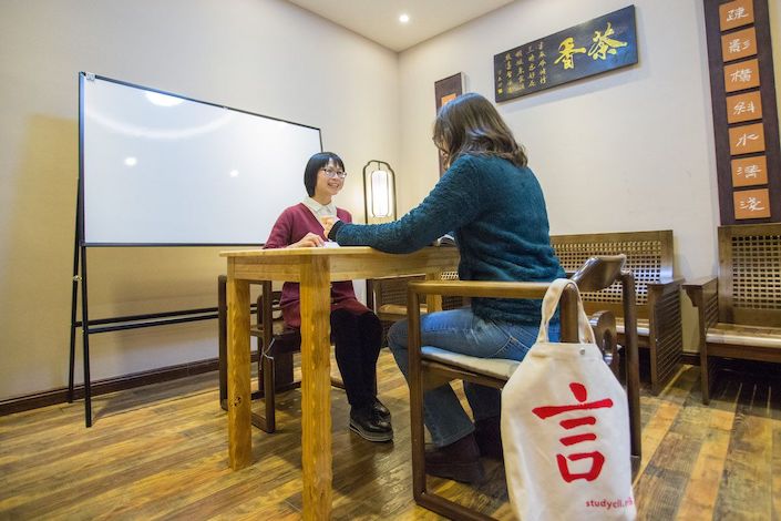 两个女人坐在一张木桌的两边，她们身后的架子上放着一块大白板，其中一张椅子上挂着一个带有 CLI 标志的帆布袋