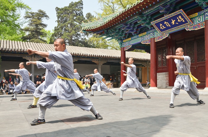 several Chinese monks in grey garments with yellow scarves around their waists practicing Chinese martial arts in front of a traditional Chinese building