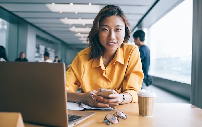 Cheerful Asian female professional in yellow blouse using smartphone and looking at camera at desk with laptop