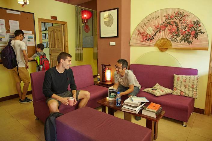 étudiant discutant avec un ami local sur des canapés violets avec un jeune garçon et enseignant face à un babillard en arrière-plan