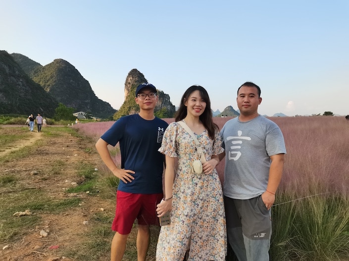 three friends standing in a field with pink flowers and a karst mountain in the background