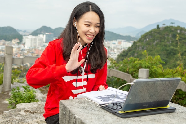 chinese teacher sitting outdoors in front of laptop computer waiving toward computer with mountains in the background
