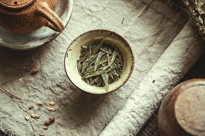 una taza de té china llena de hojas planas de té verde y una tetera parcialmente visible sobre una mesa